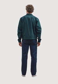 Donkergroene jas met een relaxte pasvorm, contrasterende marineblauwe broek, lichte materialen, elastische tailleband en standaard manchetten.