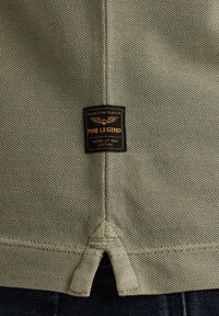 Chemise en coton kaki avec une surface texturée, présentant une étiquette noire cousue dans le bas du dos avec un texte doré et un détail d'ourlet en triangle.