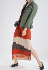 Grönt baskerjacka i militärstil över en röd mönstrad maxiklänning med krämfärgade, grå och orange ränder. Svarta ballerinaskor fulländar outfiten.