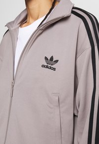 Ljusgrå zip-up-jacka med svart adidas-logotyp och ränder på axlarna. Har sidofickor och ribbad textur. Mjuk tygfinish.