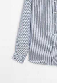 Chemise à manches longues avec des rayures verticales bleu marine et blanches, des boutons blancs sur le devant, et un poignet à rayures horizontales sur la manche.