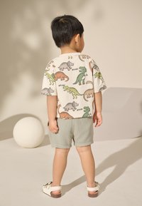 Camicia a maniche corte con motivi di dinosauri in verde, arancione e grigio su sfondo beige chiaro. Abbinata a pantaloncini di tessuto leggermente strutturato verde chiaro.