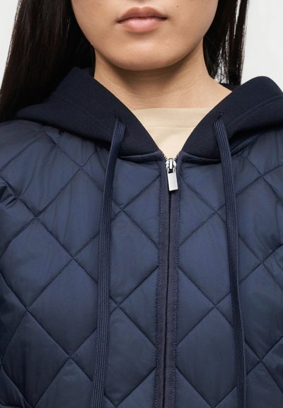 Personne portant une veste matelassée bleu marine avec une capuche et des cordons de serrage, zippée par-dessus une chemise beige.