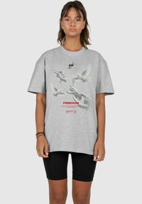 Graues, übergroßes T-Shirt aus Baumwolle mit einer Grafik von Tauben und Ketten, darunter dem Wort "FREIHEIT" in Rot.