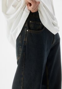 Jeans in denim blu scuro con vestibilità comoda, caratterizzati da cuciture gialle a contrasto e una piccola tasca sul retro. T-shirt bianca indossata sopra.