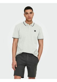 Camisa polo gris claro con ribete negro, que presenta un pequeño logotipo cuadrado negro. Combinada con pantalones cortos de color carbón, mostrando un estilo y ajuste casual.