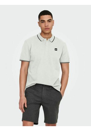 Camisa polo gris claro con ribete negro, que presenta un pequeño logotipo cuadrado negro. Combinada con pantalones cortos de color carbón, mostrando un estilo y ajuste casual.
