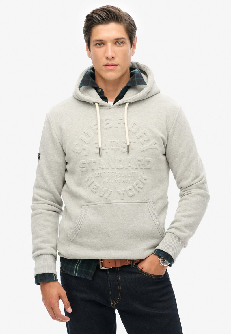 Sudadera gris claro de algodón con un bolsillo frontal, que presenta texto en relieve. Combinada con una camisa a cuadros oscura por debajo.