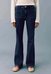 Tummansiniset leveälahkeiset farkut, joissa on korkea vyötärö, suuret etutaskut ja napitus. Yhdistetty vaalean neulepaidin kanssa.