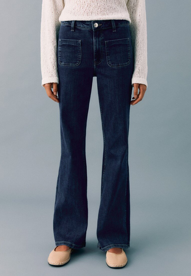 Tummansiniset leveälahkeiset farkut, joissa on korkea vyötärö, suuret etutaskut ja napitus. Yhdistetty vaalean neulepaidin kanssa.