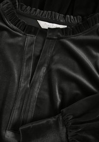Blusa di velluto nero con scollatura a V, colletto a campana e polsini arricciati. Il tessuto presenta una consistenza morbida e liscia. Etichetta del marchio visibile.