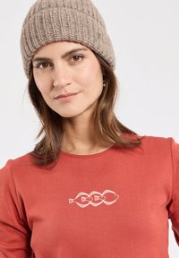 Donna che indossa un berretto di lana beige e una maglietta color ruggine con un design a catena di pesci bianchi sul petto, su uno sfondo bianco.