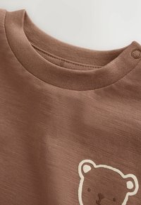 Brown baby shirt with round neckline, shoulder snap button, and simple white outline of a bear face on the chest.