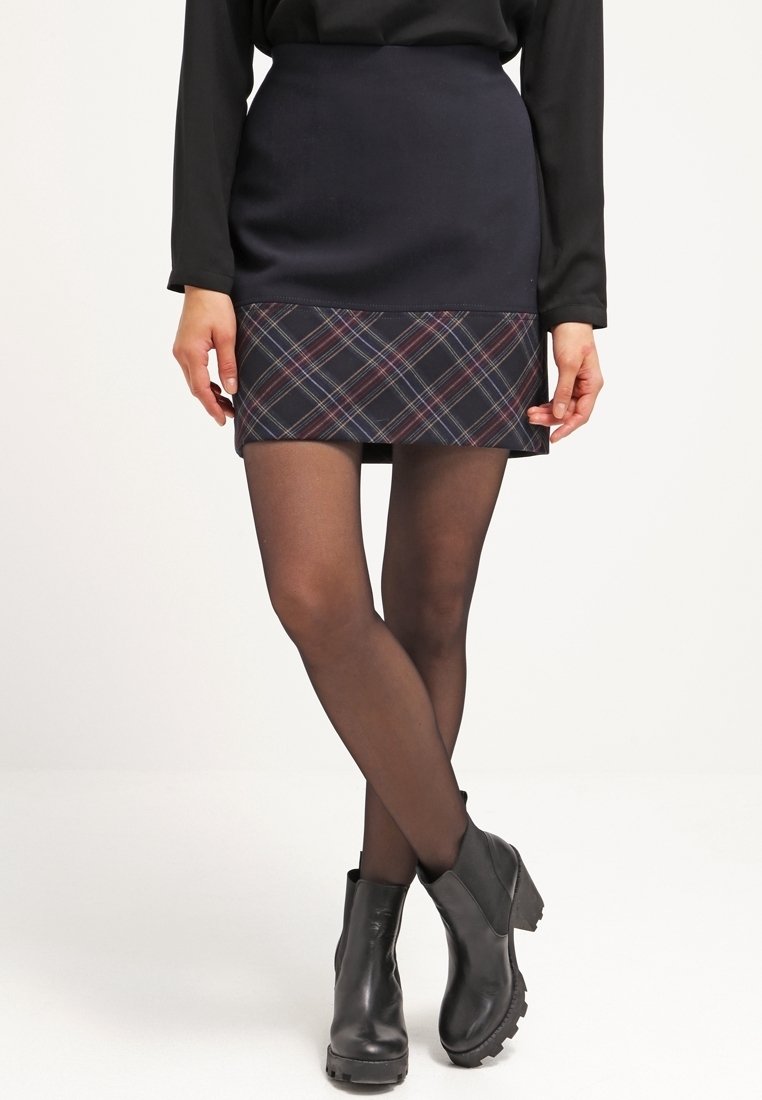 Femme portant un haut noir à manches longues, une jupe bleu marine avec un ourlet à carreaux, des collants noirs transparents et des bottines noires, debout jambes croisées.