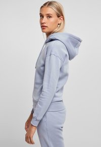 Sudadera con capucha de color azul claro hecha de una tela suave. Presenta un bolsillo tipo canguro y puños acanalados. Modelo mostrado en una pose de perfil lateral.
