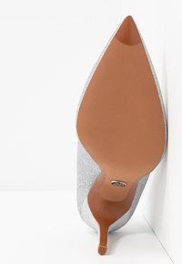 Chaussure à talon haut argenté pailleté à bout pointu vue de la semelle, montrant une semelle marron clair et un talon aiguille étroit.