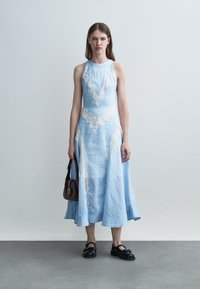 Młoda kobieta stojąca w niebieskiej sukience midi bez rękawów z białymi koronkowymi detalami, trzymająca małą ciemną torebkę i nosząca czarne buty.