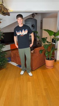 Noor mees naeratab, kandes musta graafilise trükiga T-särki ja rohelist pükse, seisab elutoas oranži diivani, suure taime ja kaunistatud kuusepuu kõrval.