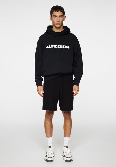 Svart hettegensere med hvitt logo, kombinert med svarte shorts. Modellen har på seg hvite sokker og sneakers, og står foran en ensfarget bakgrunn.