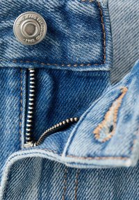 Gros plan d’un jean en denim bleu montrant une fermeture éclair métallique partiellement ouverte et un bouton gravé de « KING JEANS ESTABLISHED 1981. »