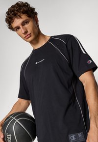 Černé tričko s krátkým rukávem a bílým lemováním, vyšívané logo šampiona na hrudi a malá nášivka na lemu; model drží basketbal.