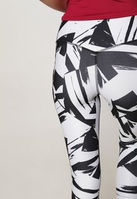 Mujer vestida con leggings de patrón abstracto en blanco y negro y una camiseta corta roja, vista desde la cintura hacia abajo, de pie contra un fondo liso.