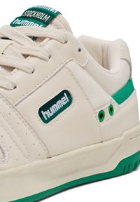Cremefarbene Sneakers mit grünen Akzenten, ausgestattet mit einem strukturierten Leder-Obermaterial, Perforationen und einem grünen Logo-Patch an der Seite.