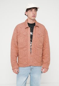 Μπουφάν σε κοραλί απόχρωση με λουστραριστή υφή, κλείσιμο με κουμπιά μπροστά, διαθέτει δύο τσέπες στο στήθος και κεντήματα με λουλουδάτο μοτίβο. Φοράται πάνω από μαύρο μπλουζάκι.