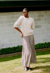 Beiger Strickpullover mit kurzen Puffärmeln, kombiniert mit einem langen, schwarz-elfenbeinfarbenen Rock mit wellenförmigem Muster und schwarzen Stiefeletten. Gras-Hintergrund.