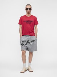 Άνδρας φοράει γυαλιά ηλίου, κόκκινο t-shirt Dsquared2 Milano, γκρι σορτς με κείμενο "ICONique", λευκές κάλτσες και μπεζ αθλητικά παπούτσια, στέκεται με τα χέρια στις τσέπες.