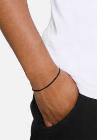 Zwarte gevlochten armband die om de pols wordt gedragen, met een eenvoudig ontwerp en een gladde textuur, tegen een witte shirtachtergrond.