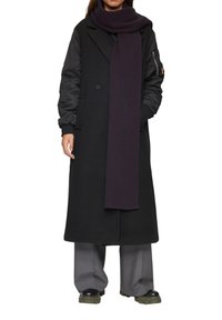 Long manteau noir avec des manches noires contrastées, écharpe violette, pantalons gris à large jambes et chaussures noires imposantes. Texture lisse et design ajusté.