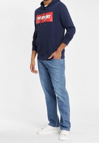 Sudadera azul marino con un logo rojo de "Wrangler", combinada con unos vaqueros de denim azul claro y zapatillas blancas, con un diseño sencillo y un corte relajado.
