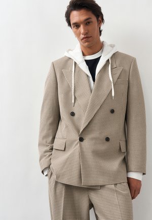 Νέος άνδρας που φοράει μπεζ καρό διπλόσαρκο κοστούμι με άσπρη φούτερ κάτω, στέκεται μπροστά σε απλό λευκό φόντο.