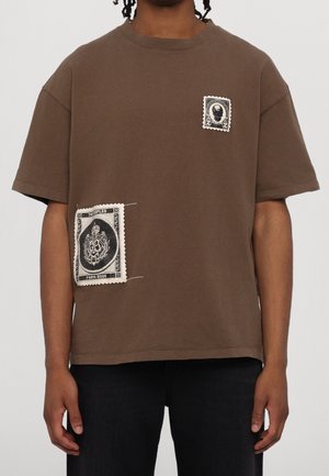 Personne portant un t-shirt marron à manches courtes avec deux patches de timbres noir et blanc, un sur la poitrine et un près de la taille.