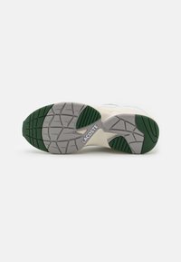 Semelle d’une basket blanche avec des motifs de bande de roulement verts et gris et le nom de la marque Lacoste visible au centre.