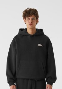 Crna dukserica napravljena od mekanog materijala, s prednjim džepom, rebrastim manžetama i malim logotipom na prsima. Relaxed fit s kapuljačom.