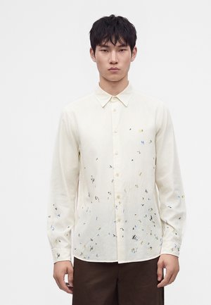 Homme portant une chemise blanche boutonnée avec de petits motifs abstraits colorés et un pantalon marron foncé sur un fond uni.