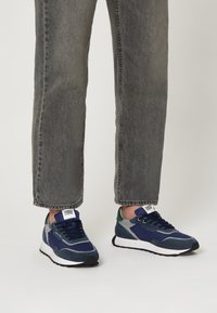 Personne portant un jean gris délavé et des baskets bleu marine avec des semelles blanches et une semelle noire, se tenant sur un fond clair.