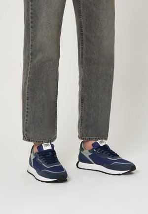 Persona che indossa dei jeans grigi sbiaditi e delle sneaker blu navy con suole bianche e battistrada nere, in piedi su uno sfondo chiaro.