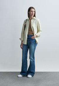 Φωτεινό κίτρινο ριγέ πουκάμισο με κουμπιά πάνω από καφέ τοπ, σε συνδυασμό με τζιν καμπάνα και λευκά αθλητικά παπούτσια. Το μοντέλο στέκεται μπροστά σε απλό φόντο.