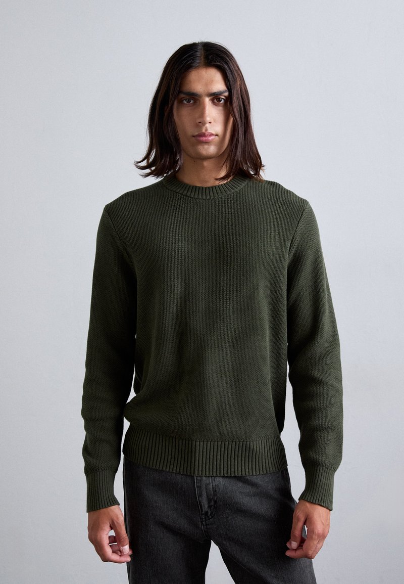 Dunkelgrüner Strickpullover mit geripptem Kragen und Bündchen, kombiniert mit dunklen Jeans. Verfügt über ein strukturiertes Muster und eine normale Passform.