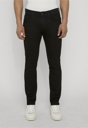 Uomo che indossa jeans neri aderenti e sneaker bianche, in piedi con le mani rilassate lungo i fianchi davanti a uno sfondo semplice.