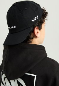 Cappello nero con visiera curva, con la scritta "VANS" ricamata in bianco sul davanti e un motivo a scacchi come accento laterale.