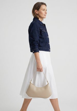 Frau trägt eine dunkelblaue Jeansjacke und einen weißen Rock, hält eine beige Handtasche mit einem einzelnen Riemen und geht vor einem schlichten Hintergrund.