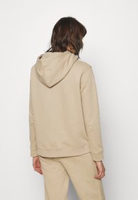 Beżowa bluza z kapturem wykonana z miękkiego materiału, charakteryzująca się luźnym fasonem, ściągaczami przy mankietach oraz kieszenią kangurką. Zaprezentowany widok z tyłu.