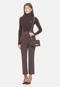 Maglione a collo alto bordeaux a coste, sagomato con una tasca sul petto. Pantaloni marroni a vita alta con dettagli con zip, abbinati a una borsa coordinata e tacchi alti.