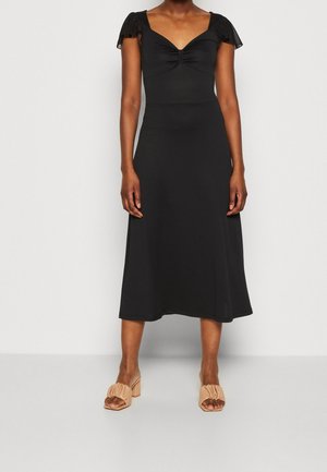 Woman wearing a black midi dress with ruched neckline and short puff sleeves, paired with beige block-heeled sandals with ruffle detail.