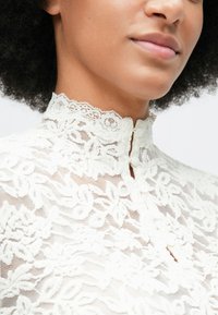 Primer plano de una persona que lleva una blusa de encaje blanca de cuello alto con patrones florales y pequeños botones a lo largo del cuello.