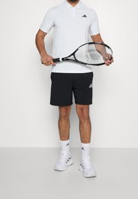 Polo blanc avec accents noirs, shorts de sport noirs, chaussures de tennis blanches et chaussettes blanches, tenant une raquette de tennis noire.